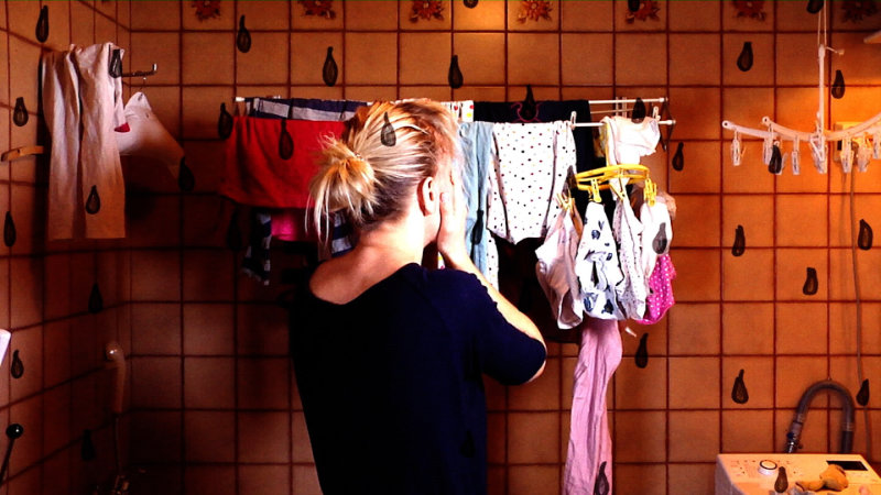 Stillbild från filmen "En arg mors bekännelser". En kvinna med händerna framför ansikte står framför en ställning med nytvättade kläder. Fotot: Impressio films.